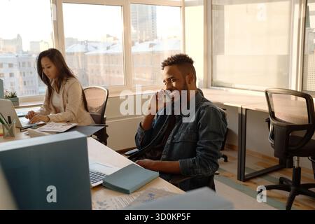 Young Asian woman working on laptop in cozy office while her black male colleague talking on smartphone and smiling Stock Photo
