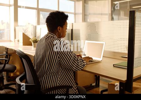 Side view of stylish African American female in striped black and white blouse working on laptop in modern office illuminated by evening sunlight Stock Photo