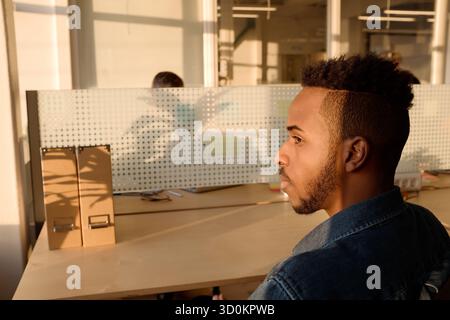 Profile portrait of young African American man in denim shirt sitting at office table and watching sunset thoughtfully Stock Photo