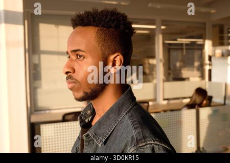 Profile portrait of thoughtful African American office worker looking through the window, his face illuminated by evening sunlight Stock Photo