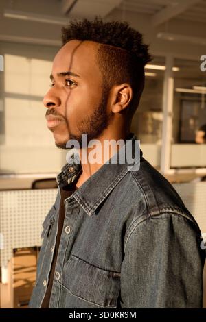 Portrait of young male African American employee standing in office and looking through the window pensively, his face illuminated by evening sunlight Stock Photo