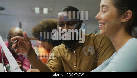 Gesturing man in brown shirt pointing at printed charts in office, with digital graphs and globe Stock Photo