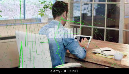 Sitting man wearing denim shirt using tablet at wooden desk, with green data graphics overlay Stock Photo