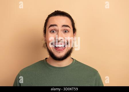 Smiling young man with beard in green shirt stands against beige background expressing joy and casual fashion suitable for lifestyle advertising Stock Photo