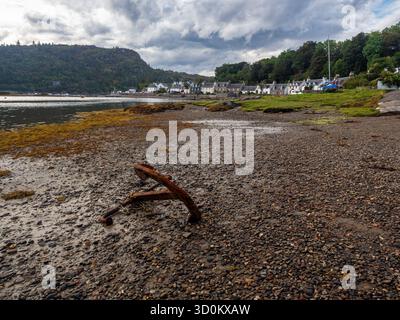 Rusty anchor on pebbled foreshore with Plockton village and hills beyond Stock Photo