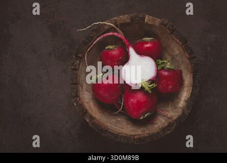A collection of vibrant red radishes sits in a carved wooden bowl on a dark, textured surface. The fresh vegetables are perfect for salads or garnishe Stock Photo