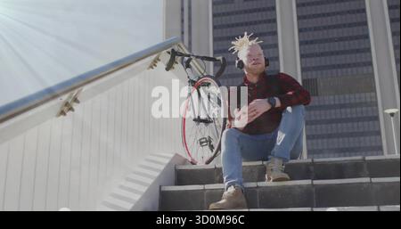 Holding smartphone, man wearing shirt, headphones sitting on urban steps by red bike, copy space Stock Photo