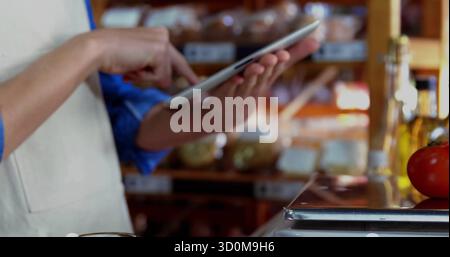 Pointing aproned worker using tablet at deli counter, with scale tomato bottles shelves, copy space Stock Photo