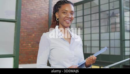 Smiling woman wearing gold hoops, holding blue folder while standing inside office near brick wall Stock Photo