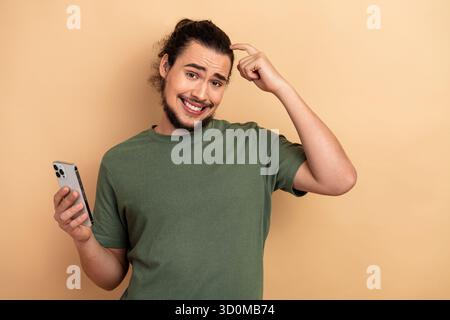 Young man with smartphone smiles and points to head in casual green t shirt against beige background Stock Photo