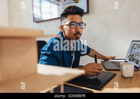Young architect working on a digital tablet at his desk, surrounded by creativity and work materials, illustrating the concept of dedication and profe Stock Photo