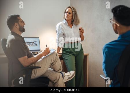 A woman in her forties in professional attire engages in discussion with two male colleagues during a team meeting at a modern architecture studio, fo Stock Photo