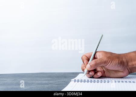 Close-up photo of a man's hand writing with a pen in a spiral notebook on a grey desk. Clean white background provides ample copy space. Stock Photo