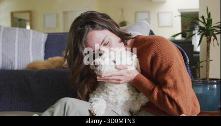 Kneeling woman holding, kissing white fluffy dog in living room with grey sofa and blue planter Stock Photo