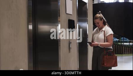 Professional in business attire checking smartphone near elevator in office lobby with tote bag Stock Photo