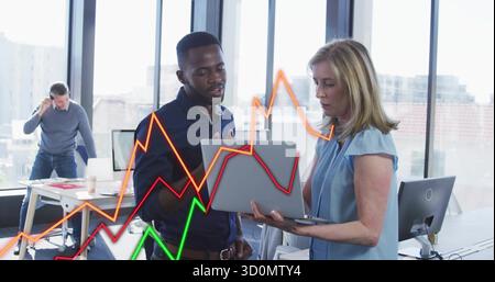 Reviewing two colleagues on laptop analyzing data in office space, with colored line chart overlays Stock Photo