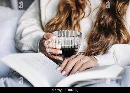 Woman holds hot coffee. Cozy morning reading. Warm sweater and long hair. Glass mug in hands. Calm home lifestyle. Winter relaxation indoors. Stock Photo