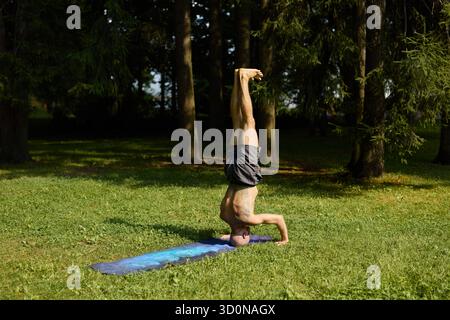 A man is practicing yoga outdoors in a headstand pose surrounded by nature and greenery Stock Photo