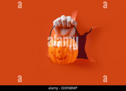 Hand Holding Jack-O'-Lantern Bucket. Hello Halloween. A hand reaches through torn orange paper holding a plastic Jack-O'-Lantern candy bucket. Stock Photo