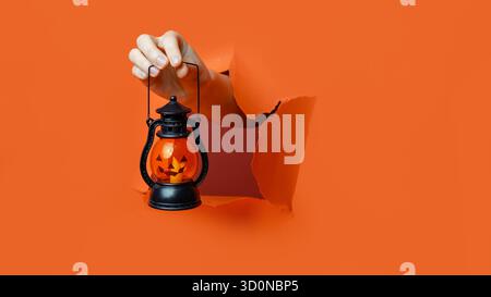 A hand holds a Halloween lantern with glowing Jack-O'-Lantern face through a ripped orange paper wall. Stock Photo