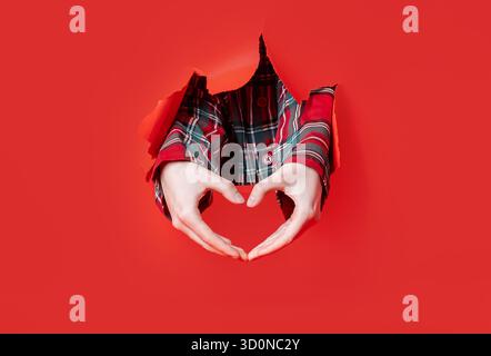 Merry Christmas. Two hands in plaid sleeves create a heart shape gesture through a torn red paper backdrop. Stock Photo