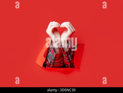 Merry Christmas. Two hands in plaid sleeves create a heart shape gesture through a torn red paper backdrop. Stock Photo