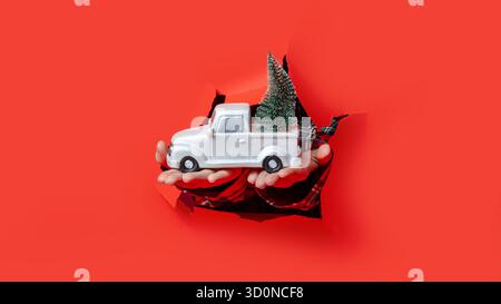 Merry Christmas. Hands in plaid sleeves hold white toy truck carrying a mini Christmas tree through a torn red background. Stock Photo