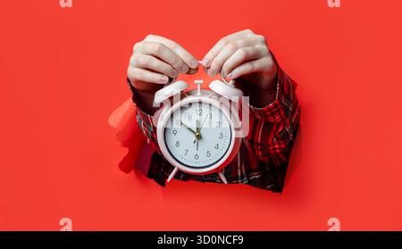 Merry Christmas. A classic white alarm clock is held in hands wearing plaid sleeves, emerging from torn red paper background. Stock Photo