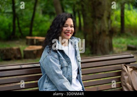 Outdoor portrait of smiling woman with natural curls. Vibrant headshot of Caucasian female with dark, curly hair, smiling directly at camera. High qua Stock Photo