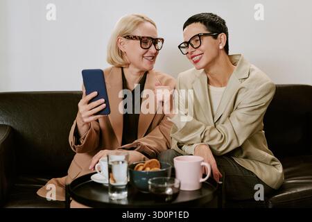 Middle Aged Women Smiling and Chatting While Using Smartphone Indoors Stock Photo