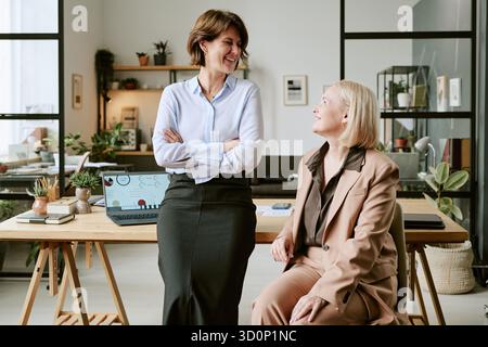 Two Middle Aged Women Smiling and Talking in Modern Office Workspace Stock Photo