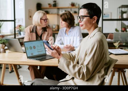 Middle Aged Woman Using Smartphone While Colleagues Discussing Project Stock Photo