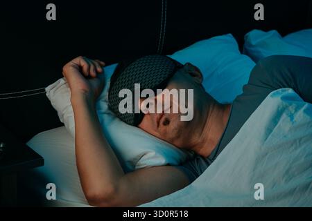 with a sleep mask covering his eyes, a man rests on his side in a bedroom, head on a pillow and body under a bed sheet, one hand curled beside his fac Stock Photo