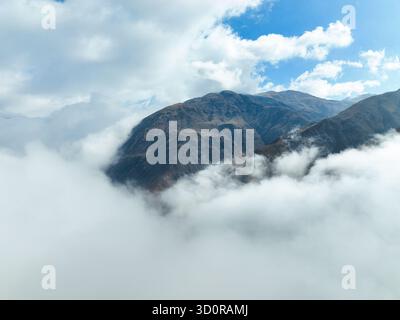 High aerial view of a rugged, dark mountain peak emerging from a thick, swirling layer of white clouds or fog under a bright, patchy blue sky Stock Photo