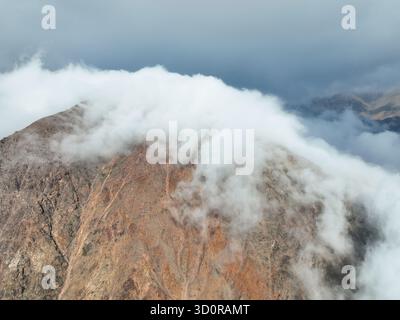 Extreme close-up aerial of a mountain's rugged, textured, reddish-brown peak with a thick layer of white clouds rolling and spilling over the summit Stock Photo