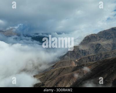 Close aerial view of a dark, steep mountain slope partially shrouded in a thick layer of white clouds under a moody, overcast sky Stock Photo