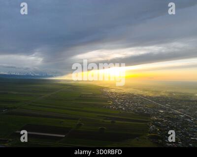 Wide aerial panoramic of patterned green fields and a long stretch of town under a dynamic sky, with the sun rising over the hazy landscape Stock Photo