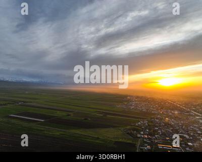 Wide aerial view capturing a dramatic split sky, with the sun setting over a sprawling suburban area and green agricultural fields leading to hazy sno Stock Photo