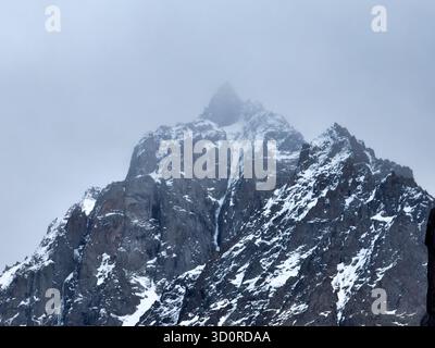 Close-up view of a rugged, snow-dusted mountain peak partially shrouded and obscured by thick fog and mist under an overcast sky Stock Photo