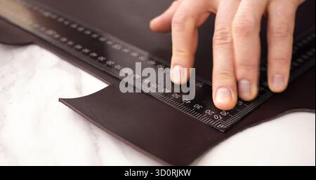 Hand measuring brown leather sheet with metal ruler on white table during crafting process Stock Photo