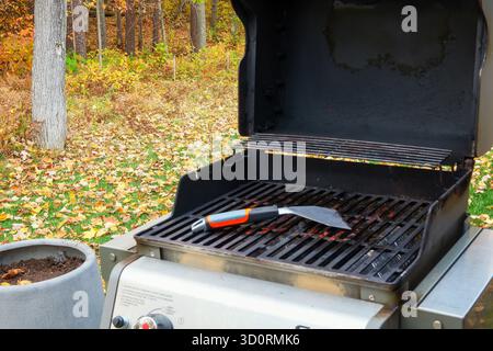 An open outdoor cooking gas grill with a metal scraper on the grates, outside in autumn with leaves on the lawn.. Stock Photo