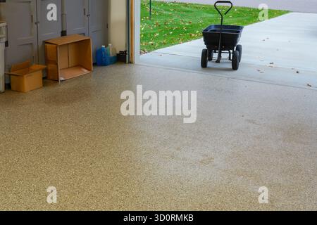 An epoxy covered garage floor with a black garden wagon cart in the open doorway near the green lawn. Stock Photo