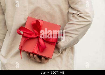 Man hiding red gift box tied with ribbon behind their back, wearing beige shirt for surprise girlfriend. Birthday present suggests surprise or special Stock Photo