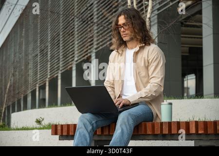 Young man, student or freelancer sits on bench using laptop for remote work at park. Outdoors. Student enjoying work break. Stock Photo