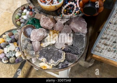 Close-up view of various rough-cut and tumbled crystals and raw stones, including rose quartz and amethyst, displayed on a metal tray for sale. Stock Photo