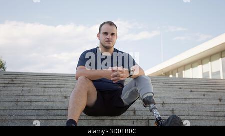 A young man sits confidently on concrete stairs, showcasing his prosthetic leg during a sunny day outdoors. Stock Photo
