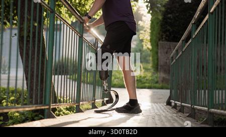 A person uses a prosthetic leg to walk across a bridge surrounded by greenery in the late afternoon sunlight. Stock Photo