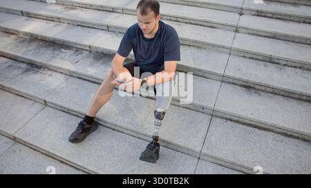 A young man with a prosthetic leg sits on stone steps, focused on his phone, enjoying a quiet moment outdoors. Stock Photo