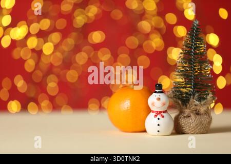 Cute decorative snowman, orange and artificial fir tree on beige table against red background with blurred lights. Space for text Stock Photo