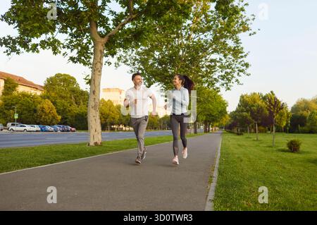 Sporty fit man and woman running together on paved street path, outdoor park trees, grass exercising Stock Photo